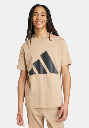 Giovane con lunghe dreadlocks che sorride, indossa una maglietta beige Adidas e pantaloni abbinati su uno sfondo neutro.