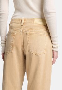 Jasnobeżowe dżinsy z denimu z tylnymi kieszeniami, delikatną teksturą i skórzaną naszywką z logo na pasku. Dopasowane w talii, luźny krój.