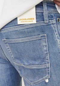Heledad sinine teksapüksid, millel on tagatasku koonusõmblusega ja silt "JACK & JONES JEANS INTELLIGENCE".