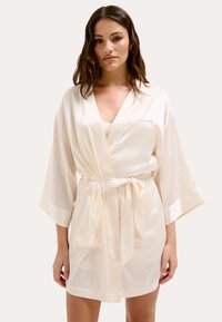 Accappatoio in satin color crema con maniche larghe, una cintura in vita e due tasche frontali; presenta un delicato ricamo sul petto.