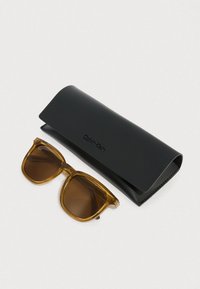 Óculos de sol Calvin Klein com lentes castanho-escuro e armação âmbar translúcida, acompanhados de um estojo preto em couro com logótipo em relevo.