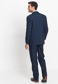Abito blu navy con un taglio sartoriale, giacca monopetto e pantaloni con singola pence. Tessuto liscio, taschino sul petto e cinque bottoni sulle maniche.