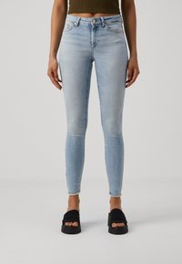 Femme portant un jean skinny bleu clair avec des ourlets effilochés et des sandales à plateforme noires à bout ouvert, debout devant un fond uni.