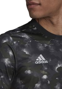 Maglietta sportiva nera a maniche corte con un motivo camouflage testurizzato, realizzata in materiale leggero, con scollatura a girocollo e logo Adidas bianco.