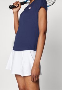 Námořnický atletický top s krátkým rukávem, v kombinaci s bílou plisovanou sukní s perforovanými detaily. Raketou na tenis drží v ruce.