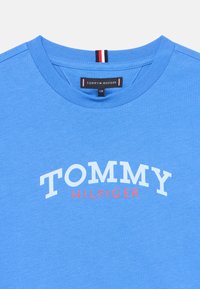 Μπλε βαμβακερό μπλουζάκι με στρογγυλή λαιμόκοψη και λευκό λογότυπο "TOMMY" με το "HILFIGER" σε κόκκινο.  Περιλαμβάνει μικρή ετικέτα λογότυπου με αντίθετες ρίγες.