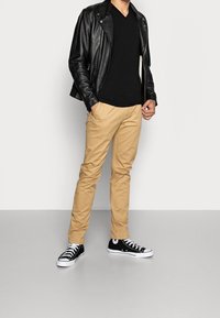 Sort læder bikerjakke, sort V-hals skjorte, khaki chinos og sorte sneakers med hvide såler. Model står mod en ensfarvet baggrund.
