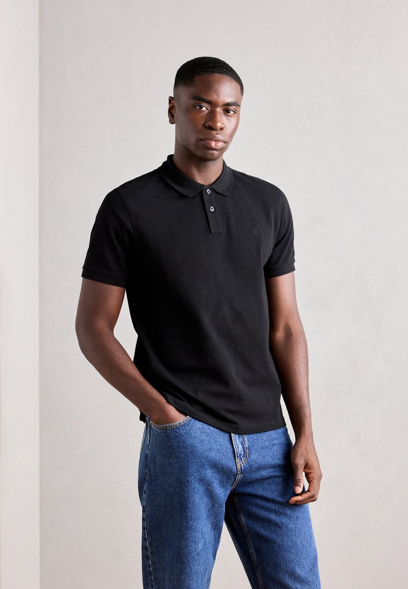 Schwarzes Poloshirt mit einer Knopfleiste mit drei Knöpfen, kurzen Ärmeln und einem dezenten Logo auf der Brust, kombiniert mit blauen Jeans.