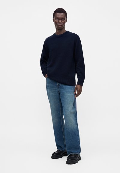 Homme debout portant un pull bleu marine foncé, un jean bleu clair et des chaussures noires épaisses sur un fond blanc uni.