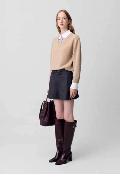 Pull en v beige clair, chemise blanche à col, shorts plissés gris charbon, bottes marron foncé jusqu'aux genoux et un sac à main assorti.