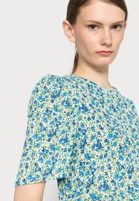 Vestido com estampa floral e mangas curtas bufantes. Fundo claro com flores azuis e verdes. Decote redondo e detalhes de botões visíveis.