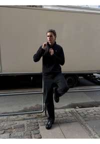 Mężczyzna w czarnej kurtce i spodniach opierający się o metalową barierkę na chodniku, trzymający zamek błyskawiczny blisko brody, w tle ciężarówka.