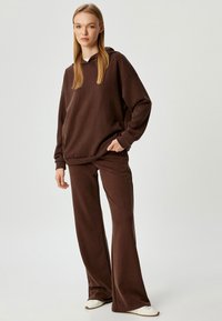 Sudadera de algodón marrón con corte relajado, bolsillo frontal y capucha con cordón, combinada con pantalones de pierna ancha a juego, que presentan una textura suave.