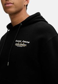 Felpa nera con cappuccio realizzata in un tessuto morbido, con un logo frontale in testo bianco e oro. Include i lacci e ha una vestibilità comoda.