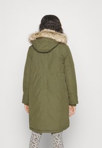 Parka verde oliva con capucha forrada de piel, mangas largas y longitud hasta la mitad del muslo. Cuenta con cintura ajustable con cordón y detalles cosidos.