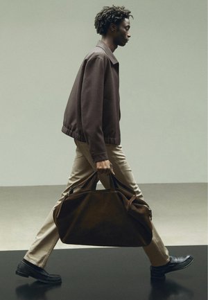 Man loopt naar rechts en draagt een grote donkerbruine sporttas, hij draagt een bruine jas, beige broek en zwarte schoenen tegen een effen achtergrond.