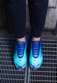 Niebieskie i turkusowe sneakersy Nike Air Max charakteryzują się falistym wzorem, teksturowanym wierzchem i ciemnoniebieskimi sznurówkami, umieszczone na metalowej siatce.