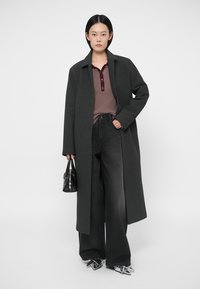 Langer schwarzer Mantel über einem bordeauxroten, gerippten Poloshirt mit Knopfleiste, kombiniert mit weiten schwarzen Jeans und metallicspitzen Stöckelschuhen. Schwarze Handtasche.