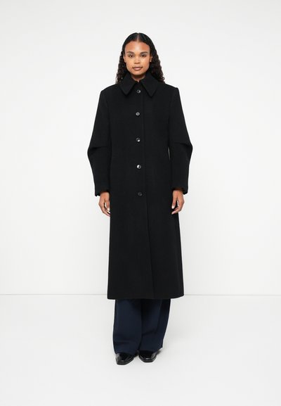 Long manteau en laine noir avec un col pointu, cinq grands boutons et des manches larges, présenté sur un fond blanc.