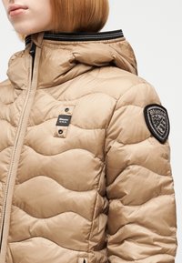 Γυναίκα φορώντας μια μπεζ καπιτονέ μπουφάν με ψηλό γιακά, μαύρες και λευκές ριγέ διακοσμήσεις και ετικέτες λογότυπου στο στήθος και στο πάνω μέρος του μανικιού.