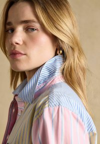 Mlada ženska z dolgimi blond lasmi nosi večbarvno pastelno črtasto srajco z dvignjenim ovratnikom in zlatimi obročastimi uhani, nevtralno ozadje.