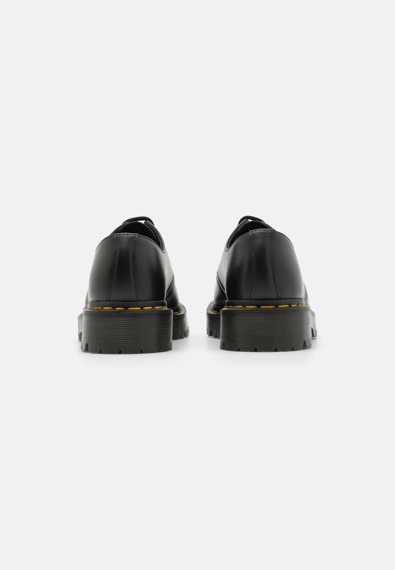 Zapatos de cuero negro con un acabado suave, suela de goma gruesa y costura amarilla a lo largo del borde. Presentan una punta redondeada y un tacón bajo.
