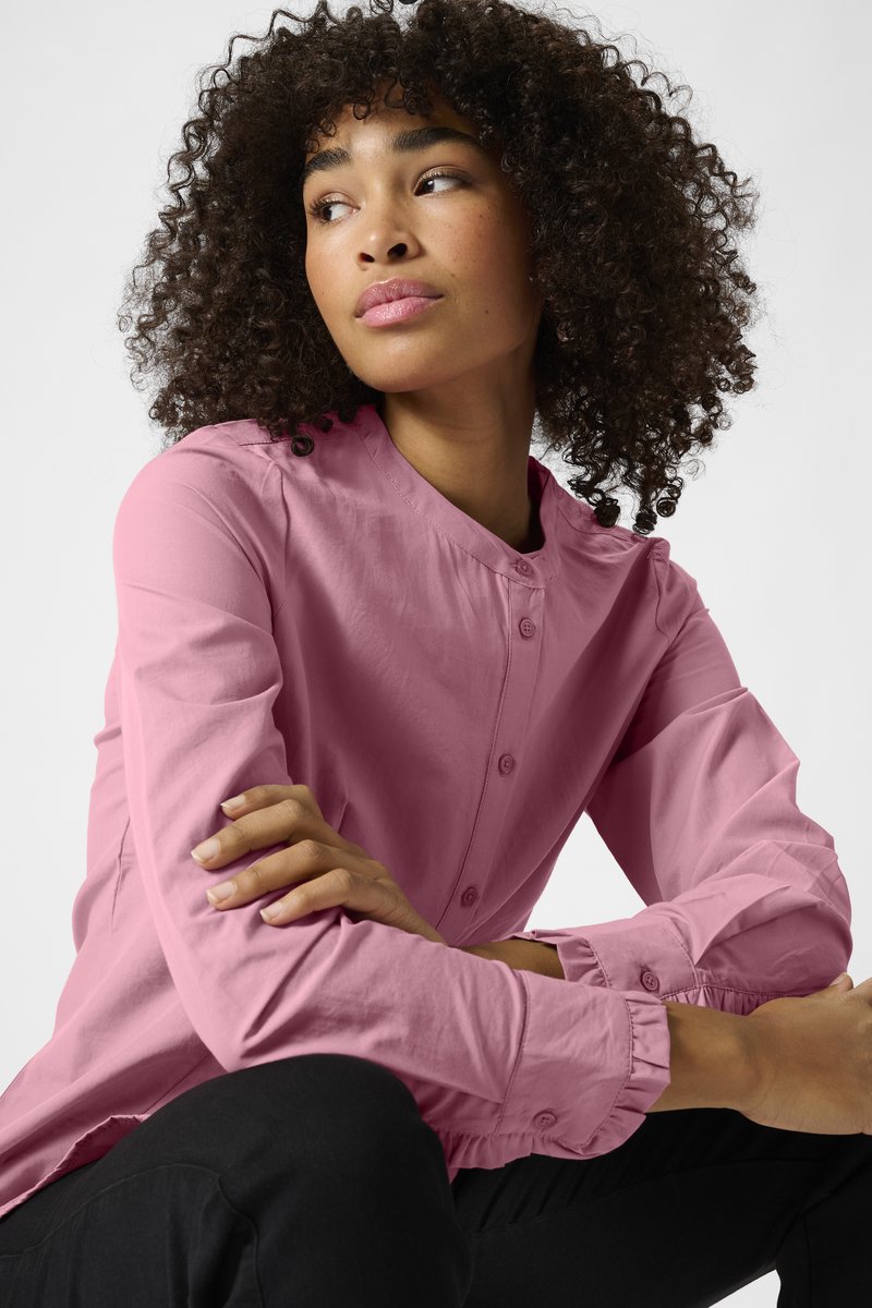 Chemise rose pâle à col rond, avec des manches longues et des poignets froncés, d'un tissu lisse.