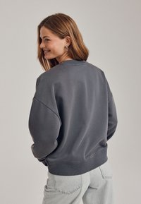 Grauer Pullover mit Rundhalsausschnitt, überschnittenen Schultern und gerippten Bündchen. Weicher Stoff mit leicht lockerem Schnitt, kombiniert mit hellgrauen Jeans.