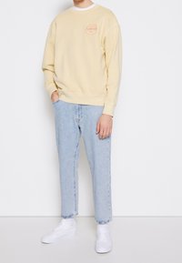 Gul sweatshirt med rund halsringning och logotyp på bröstet, i kombination med ljusblå denimjeans och vita sneakers. Enkel, avslappnad passform.