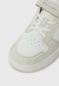 Bílé tenisky s koženým svrškem, texturovanými detaily, perforovanou špičkou a plochými tkaničkami. Mají pásek na suchý zip a gumovou podrážku.