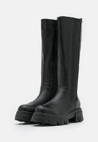 Botas negras hasta la rodilla, hechas de cuero suave, con paneles laterales elásticos, suelas de goma gruesa y punta redonda para mayor soporte y durabilidad.