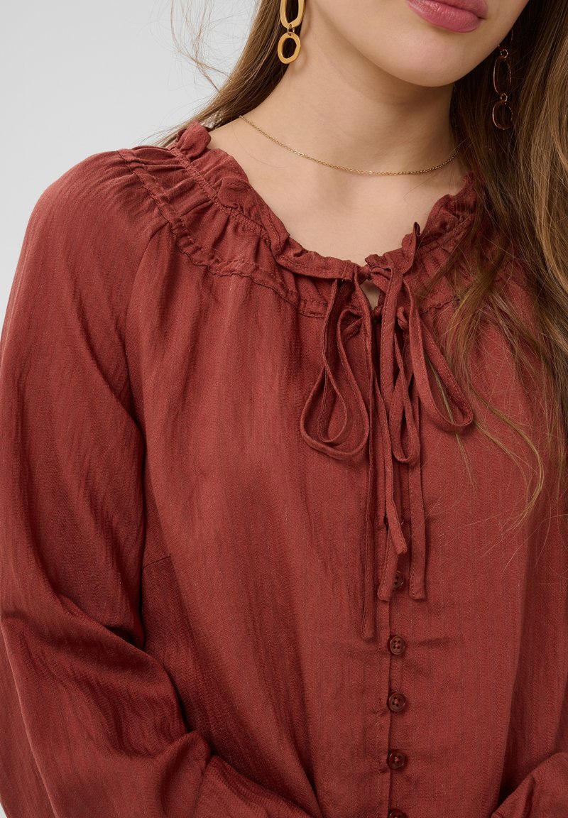 Femme portant un chemisier couleur rouille avec un col à volants noué au cou, des manches longues, un collier chaîne en or et des boucles d'oreilles géométriques en or.