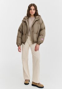 Kurtka puchowa w kolorze khaki, z teksturą w prążki, dużym kapturem, frontowymi zatrzaskami i różową metką. Połączona z kremowymi spodniami i butami.