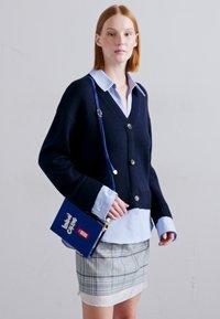 Navy knitted cardigan with large buttons, layered over a light blue shirt. Patterned pencil skirt and small blue handbag complete the outfit.