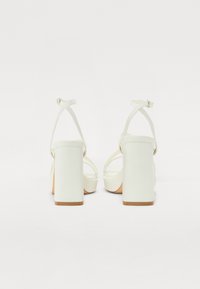 Witte blockhakken sandalen hebben dunne verstelbare enkelbandjes en een gladde, getextureerde bovenkant. De hakken zijn vierkant en iets chunky.