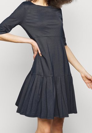 Femme portant une robe gris foncé longueur genou avec manches trois-quarts et une jupe plissée, sur un fond clair uni.