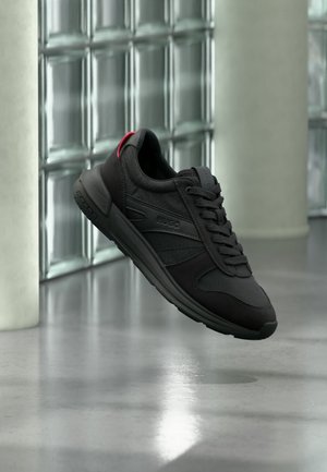 Sneaker nero Hugo con lacci e linguetta rossa sul tallone, che fluttua sopra un pavimento in cemento lucido vicino a una parete e una colonna di blocchi di vetro.