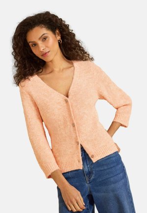 Frau mit lockigem Haar, die eine weiche pfirsichfarbene, geknöpfte Strickjacke und blaue Jeans trägt und mit einer Hand in der Hüfte vor einem weißen Hintergrund posiert.