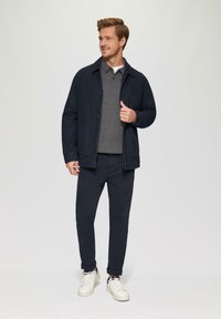 Marine knoopjasje over een grijze polo, gecombineerd met donkere broek en witte sneakers. De outfit heeft een casual pasvorm en zachte texturen.