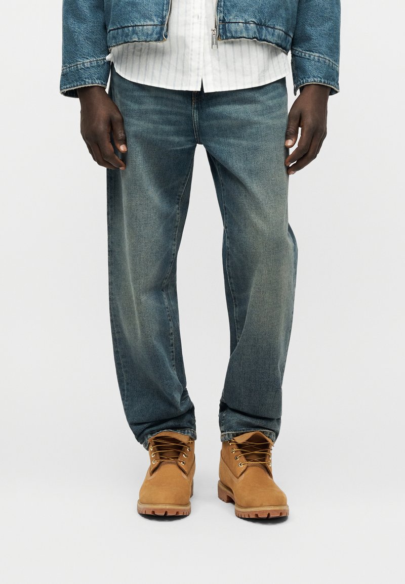 Homme portant un jean bleu délavé, des bottes de travail couleur camel, une chemise blanche à rayures et une veste en jean raccourcie, debout devant un fond uni.