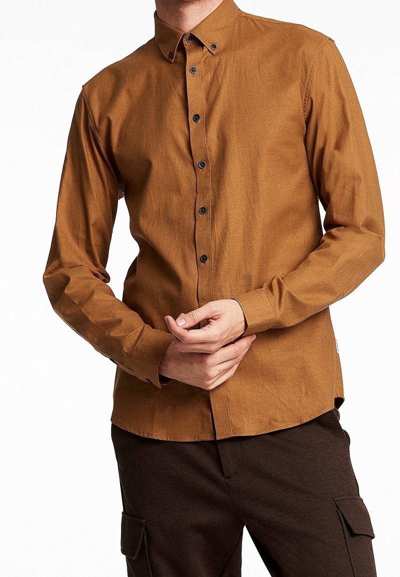 Camicia marrone a maniche lunghe con un finish testurizzato, bottoni neri. Indossata con pantaloni cargo scuri, dotati di tasche laterali.