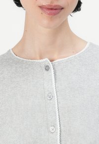 Lys grå strikket topp med rund hals, med kontrasterende hvit kant og tre knapper foran. Flat strikket tekstur.