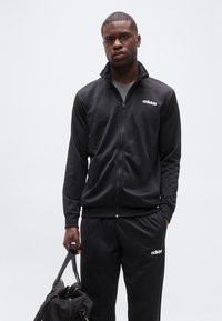 Άνδρας με μαύρη φόρμα Adidas κρατώντας μαύρη τσάντα duffel μπροστά σε απλό φωτεινό φόντο.