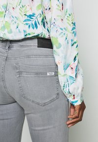 Jeans in denim grigio con una texture liscia e una vestibilità slim. Le tasche posteriori presentano dettagli di cucitura e un piccolo etichetta. La blusa floreale aggiunge un contrasto di colore.