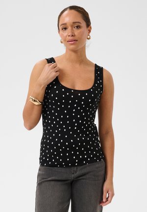 Femme aux cheveux bouclés portant un haut noir sans manches à pois blancs, des boucles d'oreilles créoles en or, un bracelet en or et un jean gris.