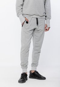 Szare dresowe spodnie z miękkiego materiału, z elastycznym paskiem w talii, zamykanymi na zamek kieszeniami i ściągaczami przy nogawkach, w połączeniu z czarnymi trampkami.