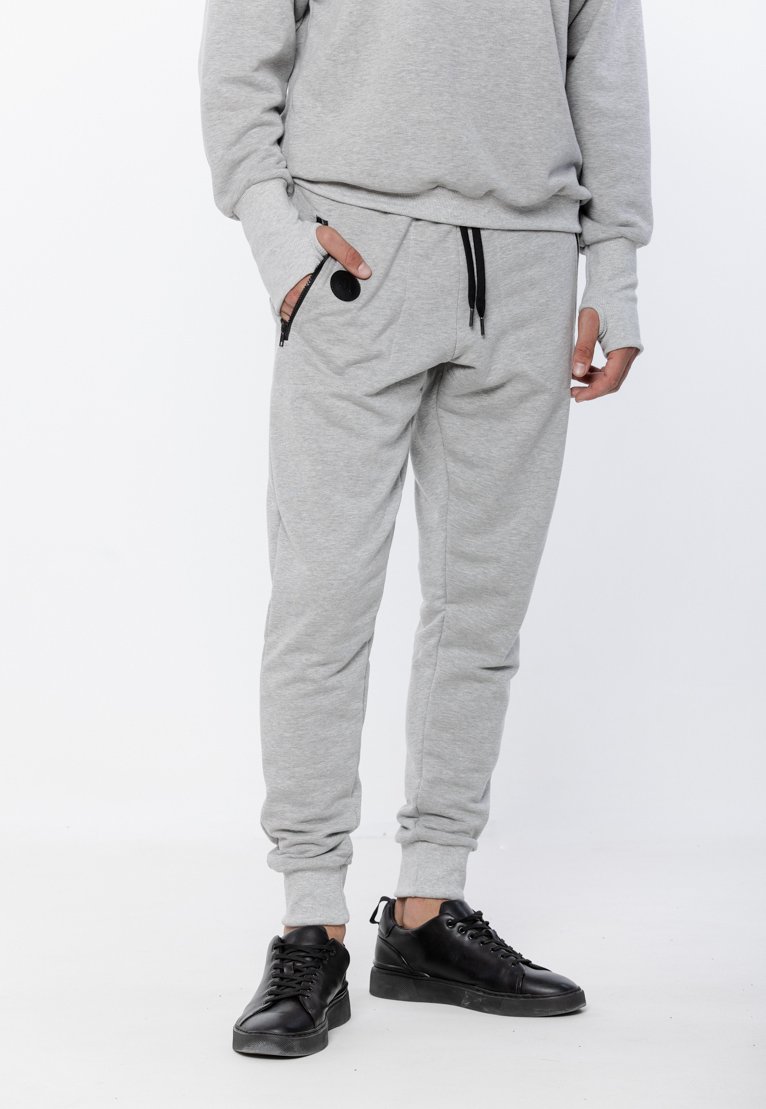Szare dresowe spodnie z miękkiego materiału, z elastycznym paskiem w talii, zamykanymi na zamek kieszeniami i ściągaczami przy nogawkach, w połączeniu z czarnymi trampkami.