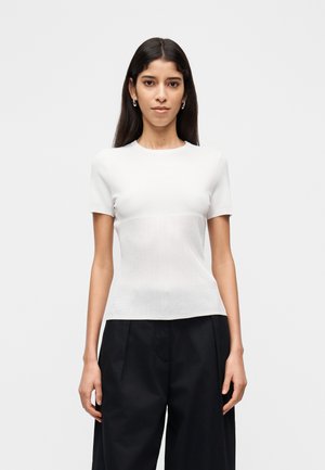 Weißes kurzärmliges Oberteil in tailliertem Design mit glatter Textur, das am Bund ein kontrastierendes, geripptes Strickmuster aufweist. Kombiniert mit schwarzen Hosen.