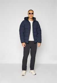 Marineblå dynejakke med hette, med horisontalt quiltet design, glidelås-lommer, båret over en hvit skjorte og svarte bukser.