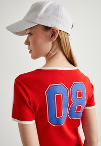 Rode katoenen t-shirt met een witte kraag, voorzien van een versleten blauwe "08" print op de achterkant. Gedragen met een witte honkbalpet en zilveren creolen.